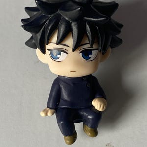 May include: A small figurine of a character with spiky black hair and blue eyes. The figure is wearing a dark blue outfit and has a neutral expression. The figurine is in a seated position with its hands resting on its knees.