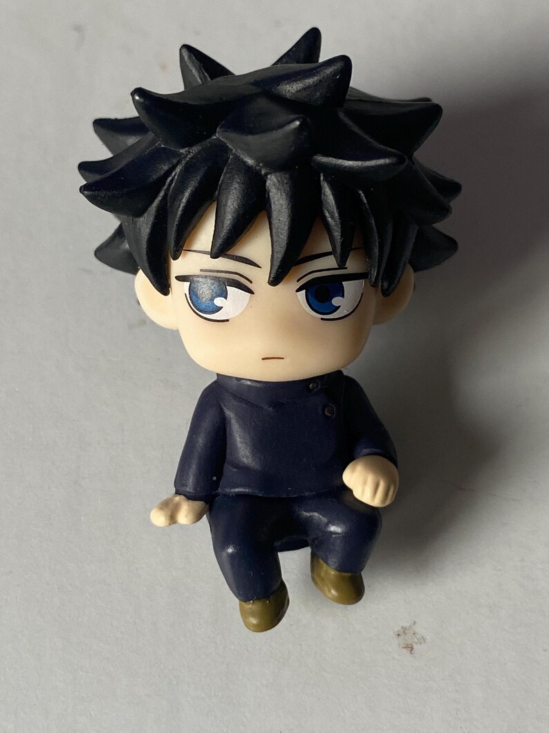 May include: A small figurine of a character with spiky black hair and blue eyes. The figure is wearing a dark blue outfit and has a neutral expression. The figurine is in a seated position with its hands resting on its knees.