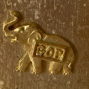 Puede incluir: Una figura de elefante dorada con las letras "GOP" en una placa rectangular. El elefante tiene colmillos visibles y una trompa enrollada. El fondo tiene un acabado dorado texturizado.