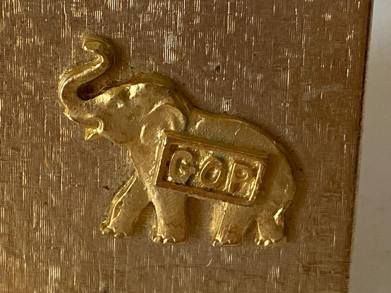 Puede incluir: Una figura de elefante dorada con las letras "GOP" en una placa rectangular. El elefante tiene colmillos visibles y una trompa enrollada. El fondo tiene un acabado dorado texturizado.
