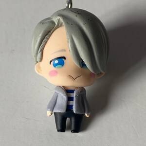 May include: A small figurine of a character with gray hair, blue eyes, and rosy cheeks. The figure wears a gray jacket over a blue and white striped shirt and black pants. A silver loop is attached to the top of the head.