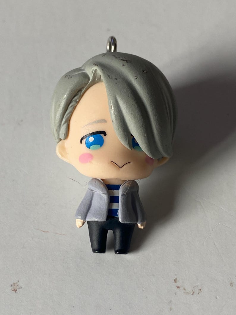 May include: A small figurine of a character with gray hair, blue eyes, and rosy cheeks. The figure wears a gray jacket over a blue and white striped shirt and black pants. A silver loop is attached to the top of the head.