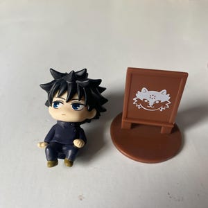 May include: A small figurine of a character with spiky black hair and blue eyes, wearing a dark blue outfit. Beside it is a brown sign with a white cat illustration.