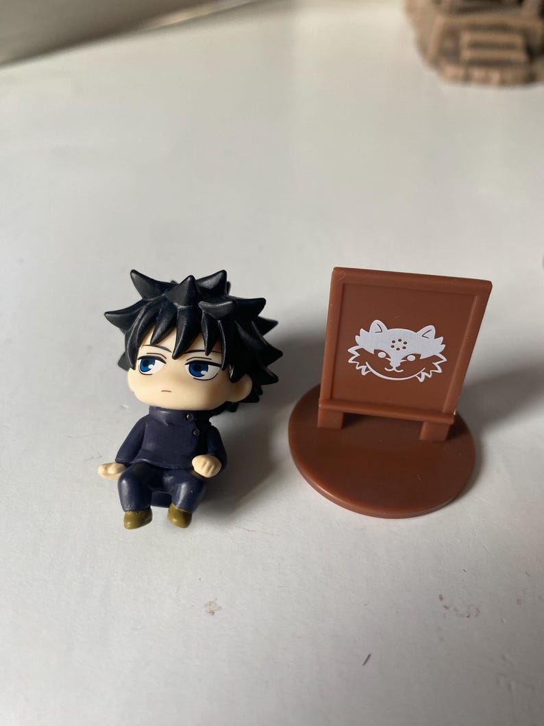 May include: A small figurine of a character with spiky black hair and blue eyes, wearing a dark blue outfit. Beside it is a brown sign with a white cat illustration.