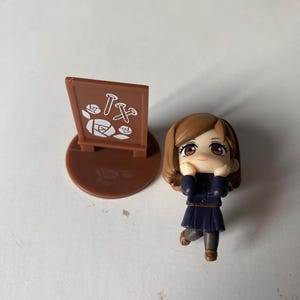 May include: A small figurine of a girl in a dark blue uniform and brown hair, with a matching brown base and a sign with white and brown graphics. The figurine is in a playful pose.