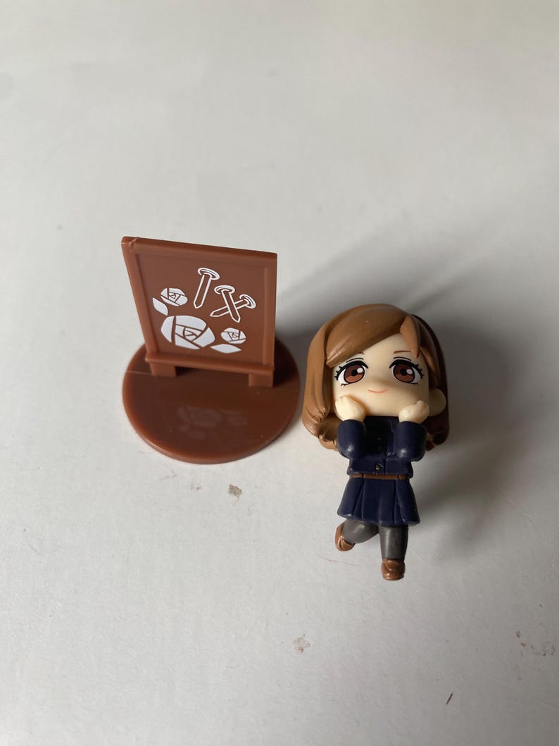 May include: A small figurine of a girl in a dark blue uniform and brown hair, with a matching brown base and a sign with white and brown graphics. The figurine is in a playful pose.