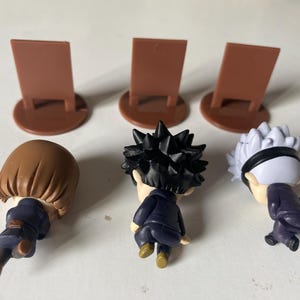 May include: Three small character figurines with brown hair, black spiky hair, and white hair with a black headband. Each figure is wearing a dark blue outfit. Three brown display stands are behind the figures.