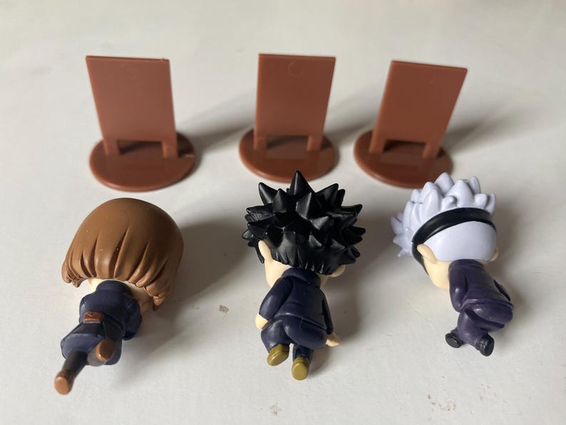 May include: Three small character figurines with brown hair, black spiky hair, and white hair with a black headband. Each figure is wearing a dark blue outfit. Three brown display stands are behind the figures.