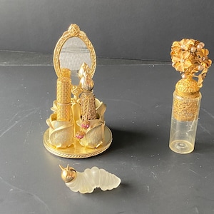 Puede incluir: Una colección de frascos de perfume antiguos y un tocador decorativo. El tocador incluye un espejo, porta lápices labiales y un frasco de perfume, todos en metal dorado. También hay un frasco de vidrio transparente con una tapa floral dorada y una oruga de vidrio.