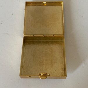 Puede incluir: Caja rectangular dorada abierta con tapa con bisagras. La caja est&aacute; hecha de un material met&aacute;lico brillante y tiene un cierre de broche. El interior de la caja tambi&eacute;n es dorado. La caja est&aacute; vac&iacute;a y se encuentra sobre una superficie clara.