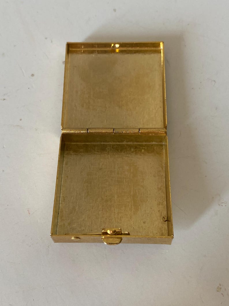 Puede incluir: Caja rectangular dorada abierta con tapa con bisagras. La caja est&aacute; hecha de un material met&aacute;lico brillante y tiene un cierre de broche. El interior de la caja tambi&eacute;n es dorado. La caja est&aacute; vac&iacute;a y se encuentra sobre una superficie clara.
