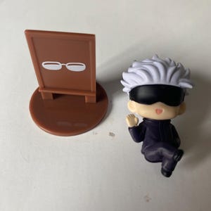 May include: A small, brown sign with a white glasses icon next to a miniature figurine of a character with white hair, a black blindfold, and a dark purple outfit. The figurine is in a seated position.