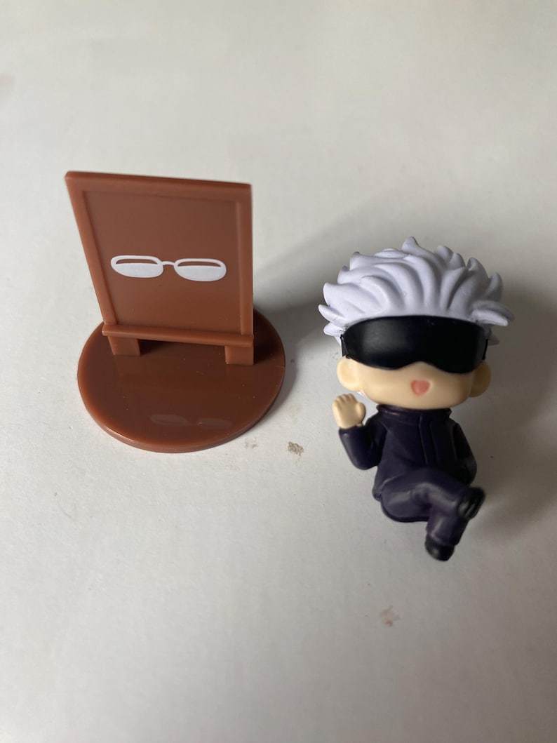 May include: A small, brown sign with a white glasses icon next to a miniature figurine of a character with white hair, a black blindfold, and a dark purple outfit. The figurine is in a seated position.