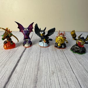 May include: Five Skylanders figures, including a purple dragon, a black bird, a gold robot, a green archer, and a black and yellow creature with a gun. The figures are all standing on small bases.