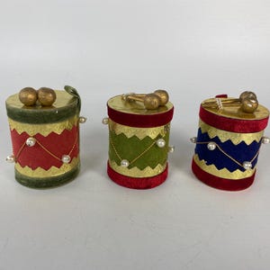 May include: Three cylindrical drum ornaments featuring velvet bodies in red, green, and blue. Each drum has gold zigzag trim, gold top surfaces with two small gold spheres, and thin gold strings adorned with tiny white faux pearls.