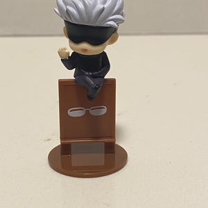May include: A small figurine of a character with white spiky hair and a black blindfold, seated on a brown stand. The stand has a pair of white glasses printed on it. The figurine is on a brown circular base.