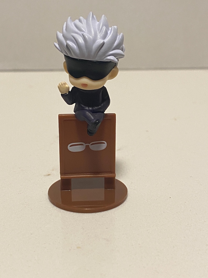 May include: A small figurine of a character with white spiky hair and a black blindfold, seated on a brown stand. The stand has a pair of white glasses printed on it. The figurine is on a brown circular base.
