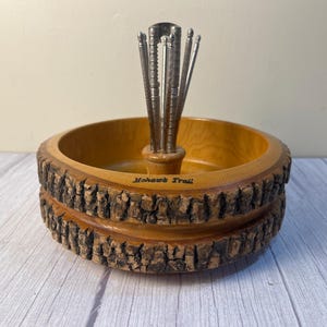 May include: A round wooden nut bowl with a natural bark exterior and a smooth interior finish. The bowl features a central metal stand holding several metal nut picks. The text Mohawk Trail is printed on the inner rim of the bowl.