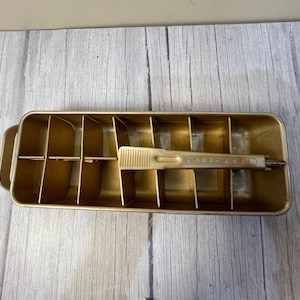 May include: A vintage gold-toned metal ice cube tray with a matching ice cube lifter. The tray is rectangular with multiple compartments. The lifter is resting inside the tray. The word "ORIGINAL" is visible on the lifter.