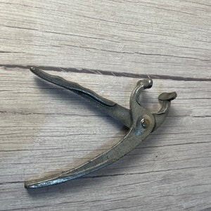 May include: A silver-colored metal tool with a textured handle and a curved design. The tool has a hinged mechanism and a rounded end, likely used for a specific task. The tool is lying on a light-colored wooden surface.