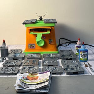 May include: An orange and green toy casting machine with multiple metal molds, bottles of glue, and a stack of instruction manuals. The molds feature various animal and object designs. The machine has a green handle and a black power cord.
