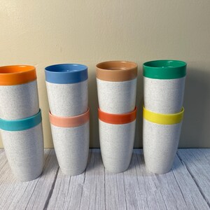 May include: Eight speckled off-white tumblers arranged in four stacks of two. Each tumbler features a solid-colored rim in orange, teal, blue, light pink, tan, red-orange, green, or yellow, creating a vibrant and colorful set of drinking cups.