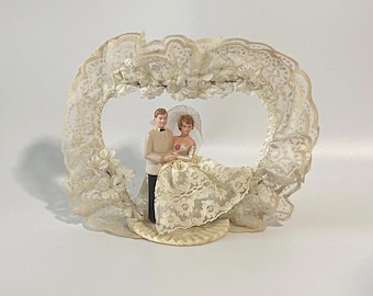 Op de afbeelding: Een vintage taarttopper voor een bruiloft met een bruid- en bruidegomfiguur. Het paar staat in een hartvormig frame versierd met kant en kleine witte bloemen. De bruid draagt een kanten jurk en sluier, en de bruidegom draagt een smoking.
