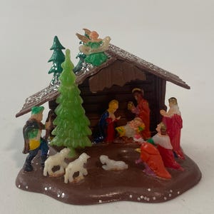 May include: A miniature plastic nativity scene featuring a brown stable with a glittery roof, a green pine tree, and an angel on top. The set includes small figures of people in colorful robes, a baby in a manger, and several white sheep.