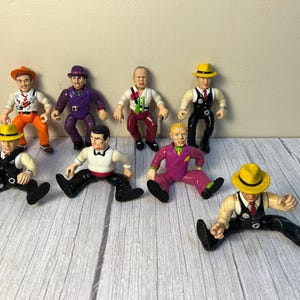 May include: A set of eight vintage action figures, all dressed in different outfits. The figures are wearing hats, suits, and ties. They are all standing on a white surface.