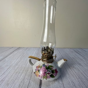 May include: A white porcelain oil lamp shaped like a teapot with a floral design. The lamp has a clear glass chimney and a gold-colored metal wick holder.