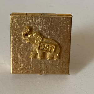 Puede incluir: Peque&ntilde;a caja dorada con superficie texturizada. Presenta un relieve en forma de elefante con las letras "GOP" en un lateral. Probablemente un art&iacute;culo de colecci&oacute;n o conmemorativo.