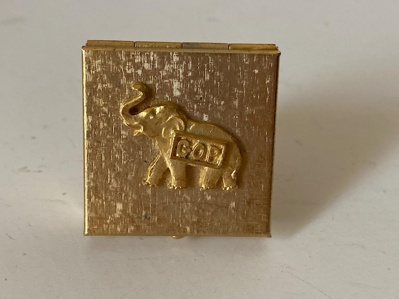 Puede incluir: Peque&ntilde;a caja dorada con superficie texturizada. Presenta un relieve en forma de elefante con las letras "GOP" en un lateral. Probablemente un art&iacute;culo de colecci&oacute;n o conmemorativo.