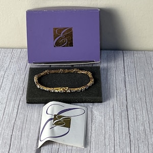May include: A gold and silver bracelet with a row of small stones. The bracelet is in a purple box with the letter "E" on it. The box is on a wooden surface.