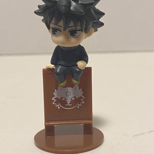 May include: A small figurine of a character with spiky black hair and blue eyes, seated on a brown sign. The sign features a white cat-like design. The figurine is on a brown circular base.