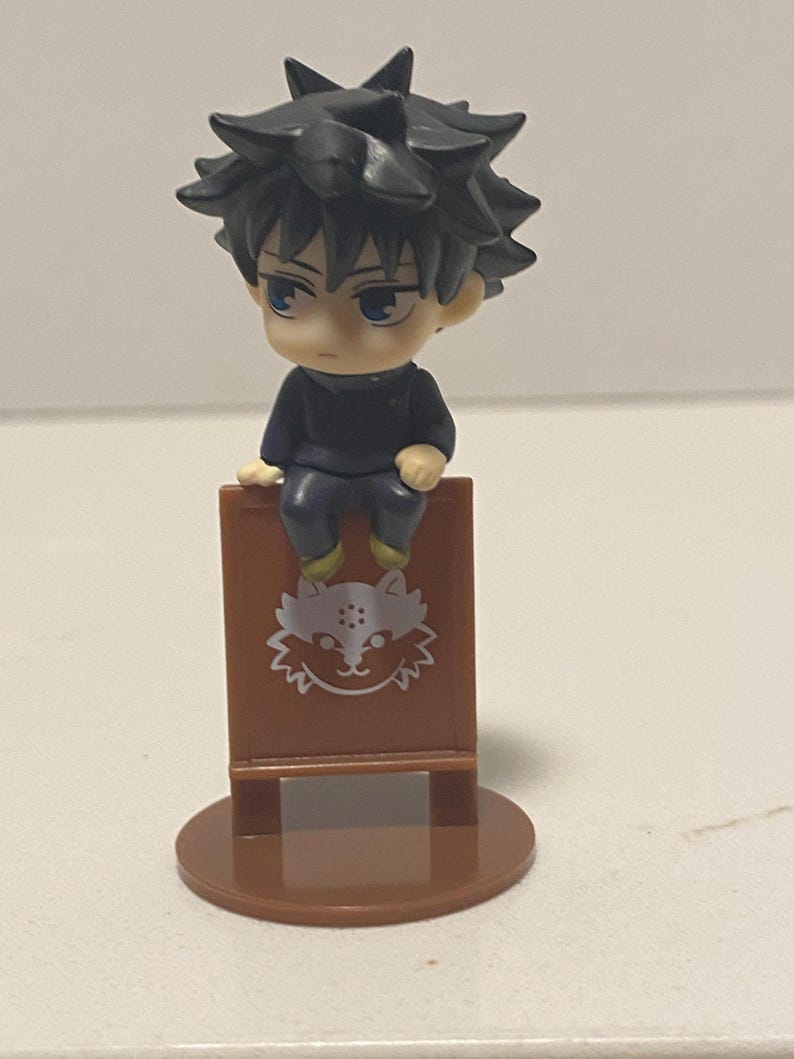 May include: A small figurine of a character with spiky black hair and blue eyes, seated on a brown sign. The sign features a white cat-like design. The figurine is on a brown circular base.