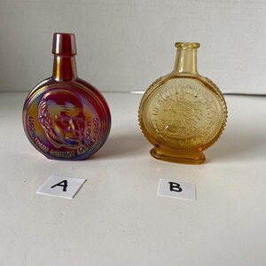 May include: Two vintage glass bottles with a round, coin-shaped design. The bottle labeled 'A' is iridescent red and features a portrait of a man. The bottle labeled 'B' is amber and features a design of a woman with a crown.