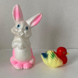 May include: A white Easter bunny figurine with pink ears and feet, and blue eyes. Beside it is a small, colorful duck figurine with a red head, yellow body, and blue and green wings. Both are likely vintage decorations.