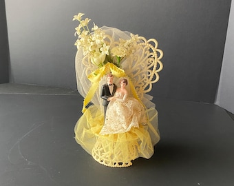 Vintage Plastic Bride & Groom Cake Topper with Yellow / White tulle, ribbon  and silk type flowers in front of Filigree heart background