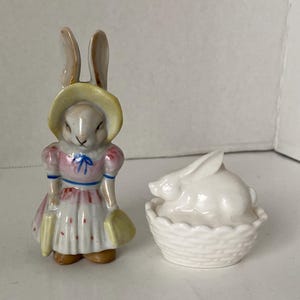 May include: Two ceramic Easter bunny figurines. One is a standing bunny wearing a pink dress and a yellow hat, holding a yellow purse. The other is a white bunny lying in a white basket. A charming Easter decoration.