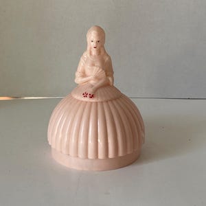 May include: A vintage, light pink figurine of a woman in a long dress, holding a fan. The dress has a pleated skirt and is adorned with small red floral accents. The figurine is likely made of plastic.