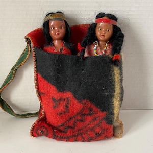 May include: Two small dolls dressed as Native Americans are inside a red and black patterned fabric pouch with a green strap. The dolls have black hair, brown eyes, and are wearing beaded necklaces.