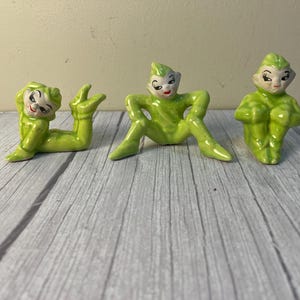 May include: Three ceramic elf figurines in a glossy lime green glaze. One elf lies on its stomach with legs raised, one sits with legs spread and hands on knees, and one sits with knees pulled up. Each figure has painted facial features.