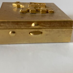 Puede incluir: Una caja rectangular dorada con un dise&ntilde;o en relieve y texturizado en la tapa. La caja tiene un cierre y un peque&ntilde;o bot&oacute;n redondeado en el lateral. El dise&ntilde;o de la tapa parece ser una representaci&oacute;n estilizada de letras o s&iacute;mbolos.