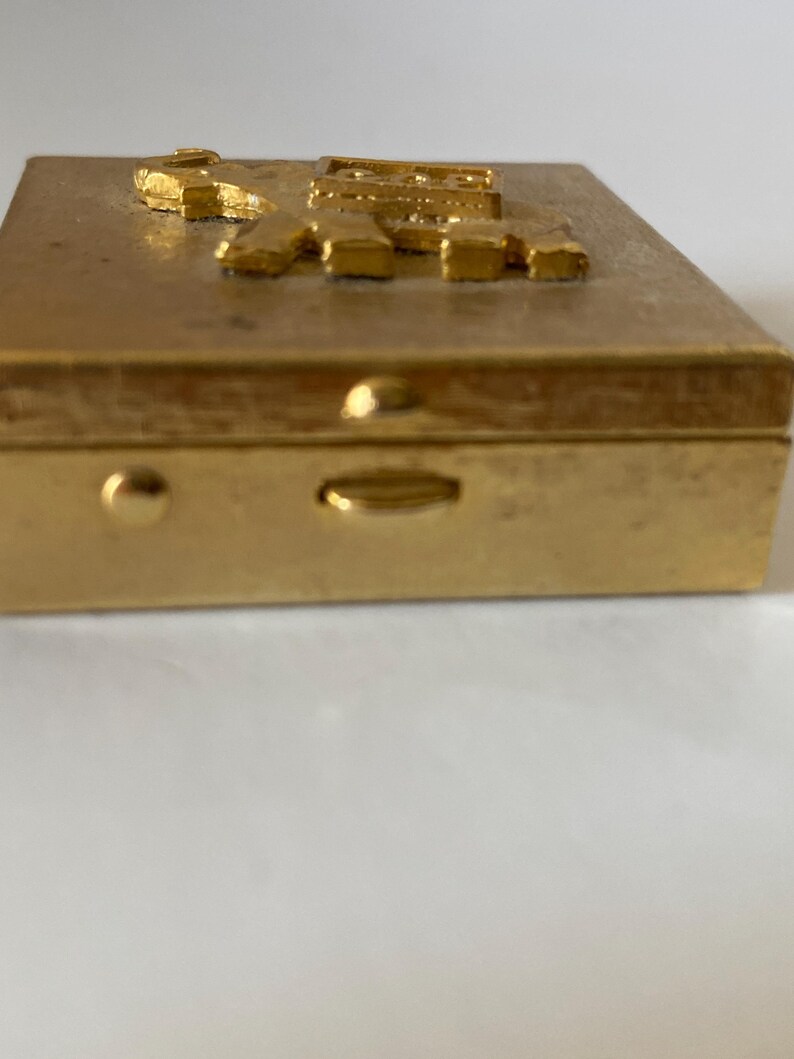 Puede incluir: Una caja rectangular dorada con un dise&ntilde;o en relieve y texturizado en la tapa. La caja tiene un cierre y un peque&ntilde;o bot&oacute;n redondeado en el lateral. El dise&ntilde;o de la tapa parece ser una representaci&oacute;n estilizada de letras o s&iacute;mbolos.