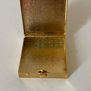Puede incluir: Caja rectangular dorada abierta con tapa con bisagras. La caja est&aacute; hecha de un metal brillante y texturizado. La tapa est&aacute; abierta, revelando el interior de la caja. La caja est&aacute; vac&iacute;a.