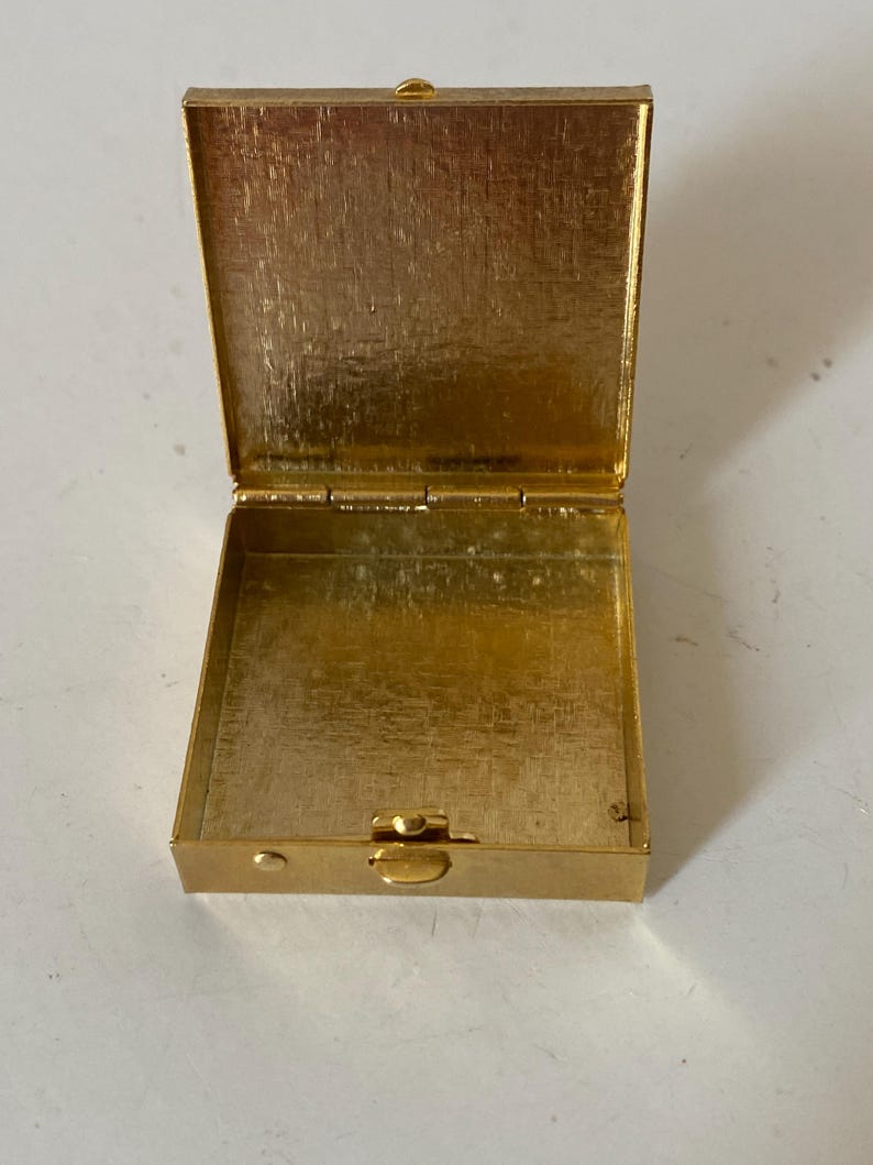 Puede incluir: Caja rectangular dorada abierta con tapa con bisagras. La caja est&aacute; hecha de un metal brillante y texturizado. La tapa est&aacute; abierta, revelando el interior de la caja. La caja est&aacute; vac&iacute;a.