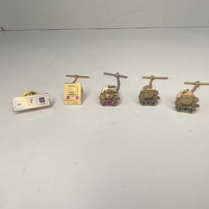 May include: A collection of gold-toned tie tacks and a pin. The tie tacks feature various designs, including a computer, a face with jewels, and a rectangular pin with a design. The tacks are adorned with colorful gemstones.
