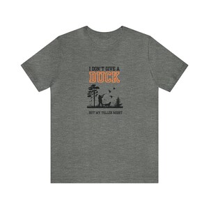 Op de afbeelding: Een heidekleurig T-shirt met een zwart silhouet van een persoon die eenden jaagt met een hond. De tekst "I don't give a duck...but my toller might" is in oranje gedrukt.