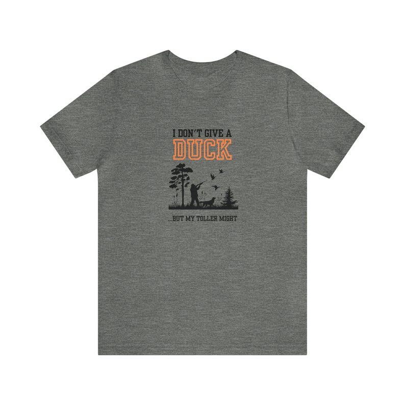 Op de afbeelding: Een heidekleurig T-shirt met een zwart silhouet van een persoon die eenden jaagt met een hond. De tekst "I don't give a duck...but my toller might" is in oranje gedrukt.