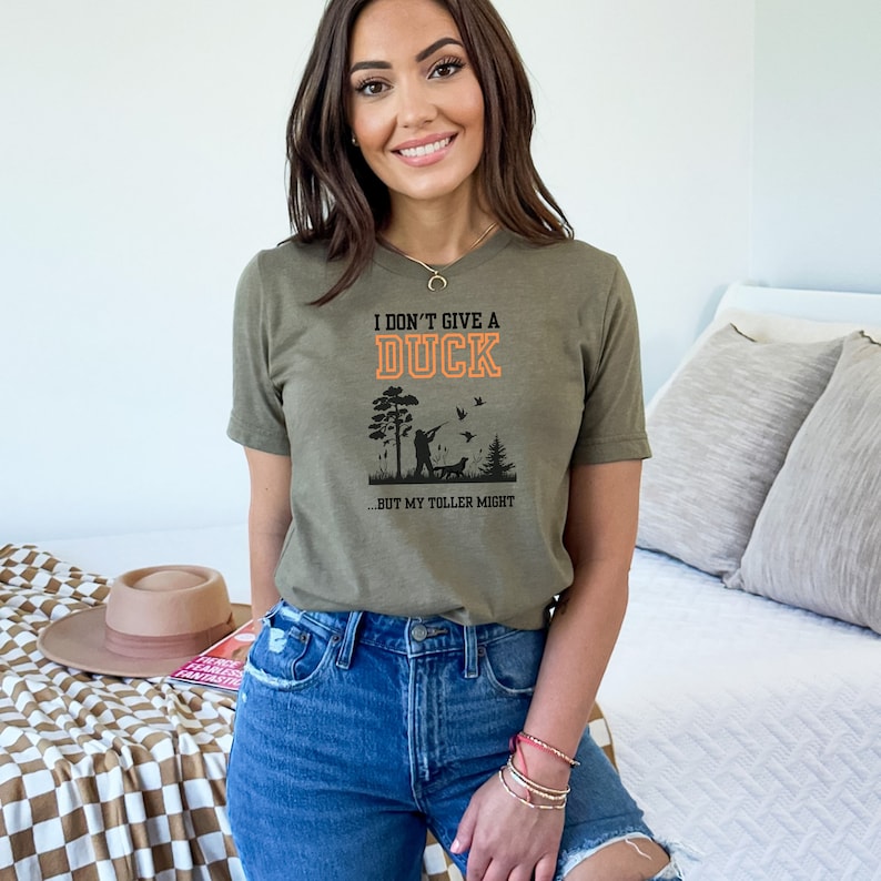 Op de afbeelding: Een groen T-shirt met een zwarte en oranje afbeelding waarop staat "Ik geef geen moer om een eend...maar mijn toller misschien". De afbeelding toont een silhouet van een persoon die eenden jaagt met een hond.